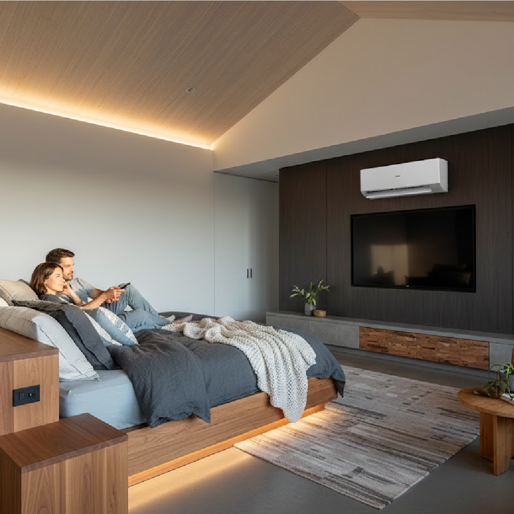 Couple relaxing on wooden platform bed in modern bedroom with dark accent wall, wall-mounted TV, white AC unit.